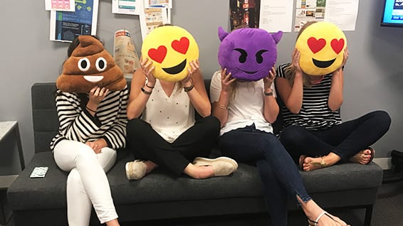 Four women sitting on couch covering their faces with emoji shaped pillows