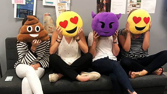 Four women sitting on couch covering their faces with emoji shaped pillows