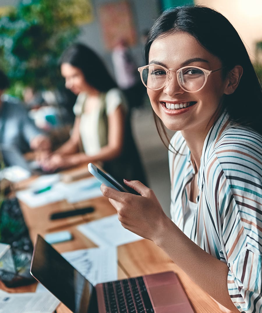 girl with glasses and laptop smiling while holding a smartphone