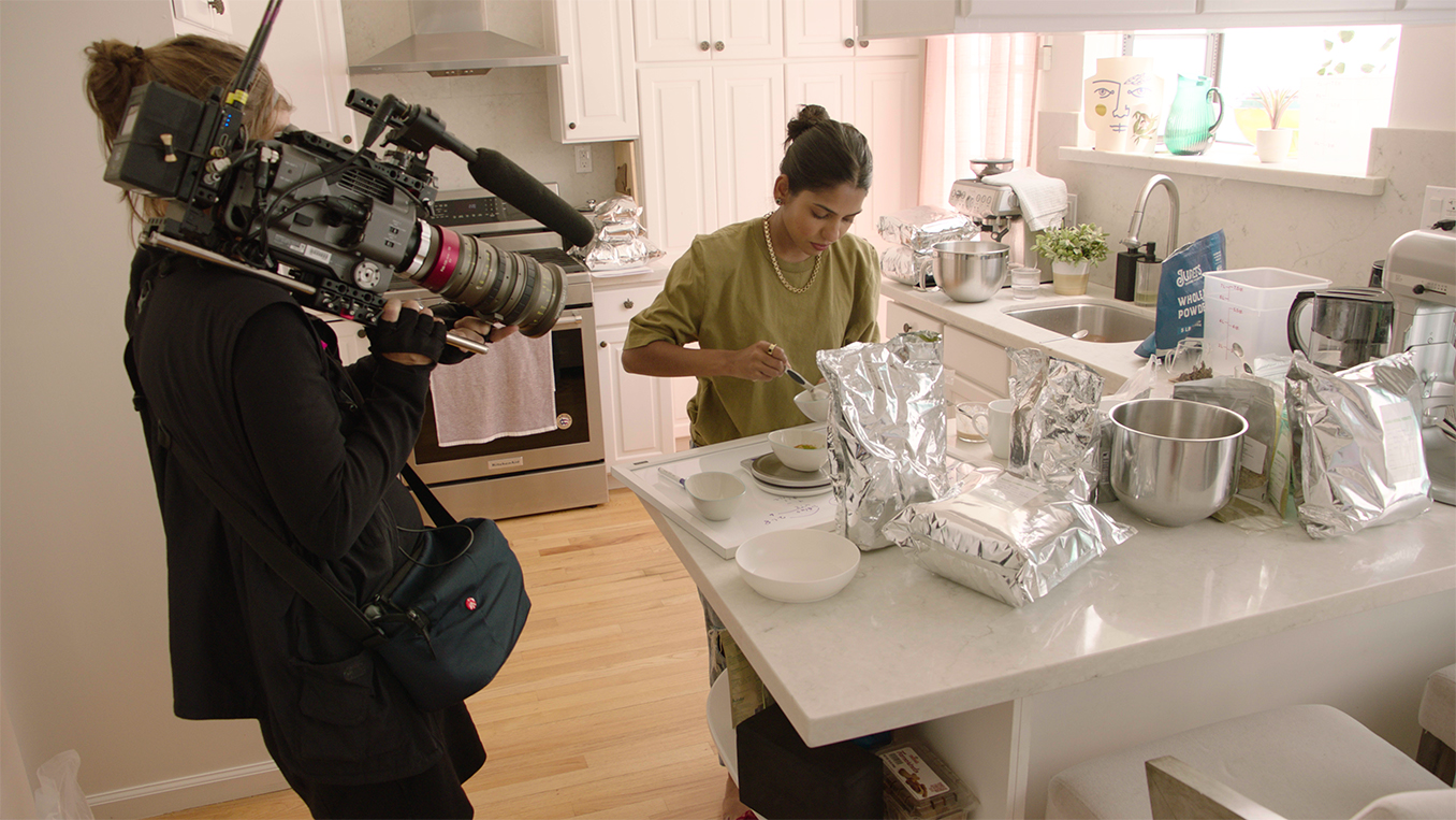 Person holding large camera filming a woman preparing food on a cutting board in a kitchen.